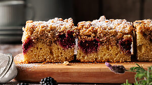 Stücke von saftiger Brombeer Streuselkuchen vom Blech in der Nahaufnahme, mit knusprigen Streuseln und deutlich sichtbarer Frangipane-Schicht auf Holzbrett angerichtet.